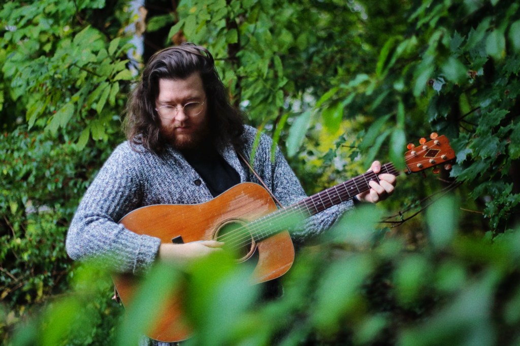 Photo of Tyler and his guitar surrounded by leaves and trees in his backyard